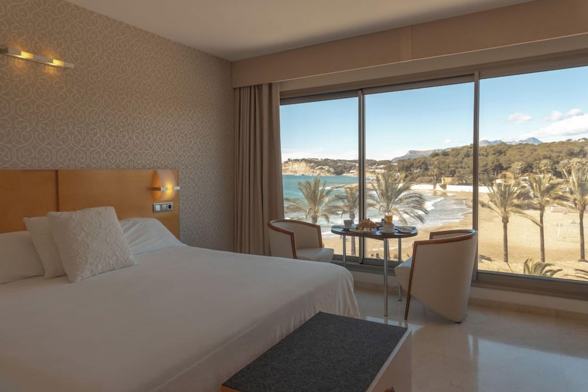 A hotel room with a large bed, a small table with chairs, and a view of a beach and mountains.