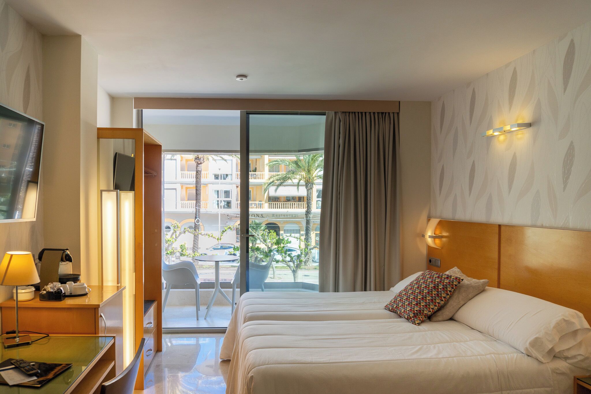 Quarto de hotel com cama, escrivaninha com luminária, televisão e varanda com vista para edifícios e palmeiras.