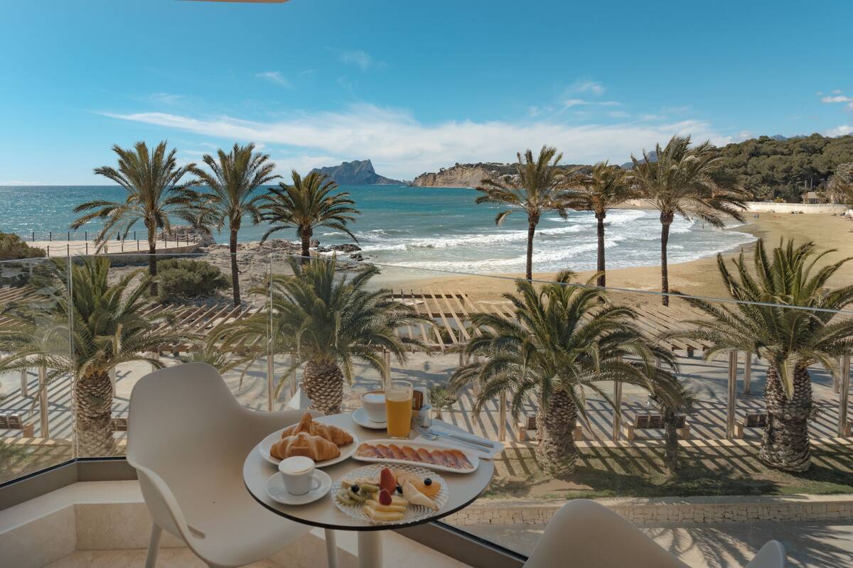 Un balcon avec une table dressée pour le petit-déjeuner, donnant sur une plage bordée de palmiers et offrant une vue sur la mer.