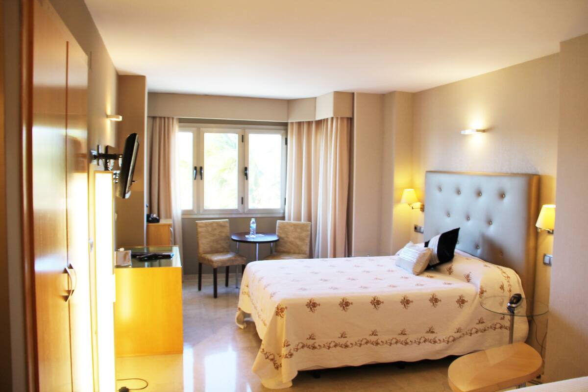 Une chambre d’hôtel avec un lit, un bureau, une chaise, une petite table et une fenêtre avec des rideaux.