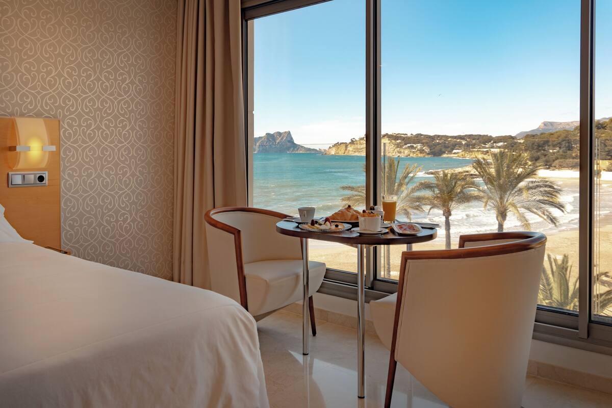 Une chambre d’hôtel avec un lit, deux chaises, une petite table avec un petit-déjeuner, et une grande fenêtre donnant sur la côte.