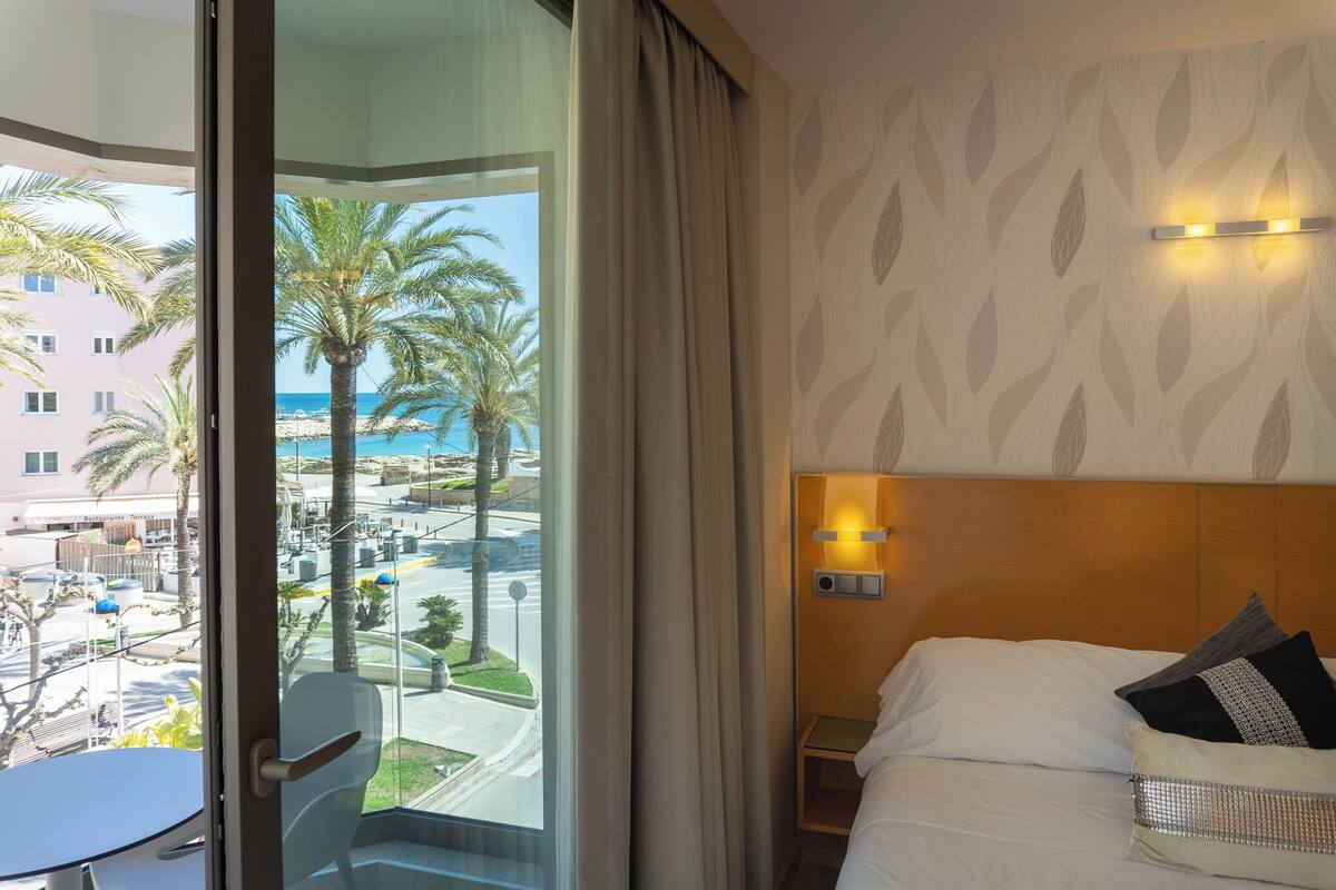 Une chambre d’hôtel avec un lit, une table de chevet, une lampe fixée au mur et une vue sur la plage et les palmiers.
