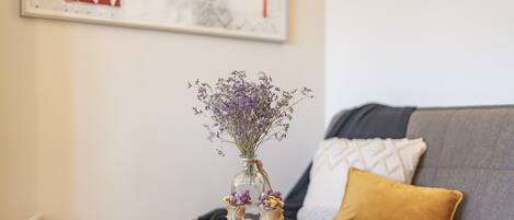 A small table with a vase of flowers and two small bottles, a grey sofa with a yellow pillow, and a wicker basket.