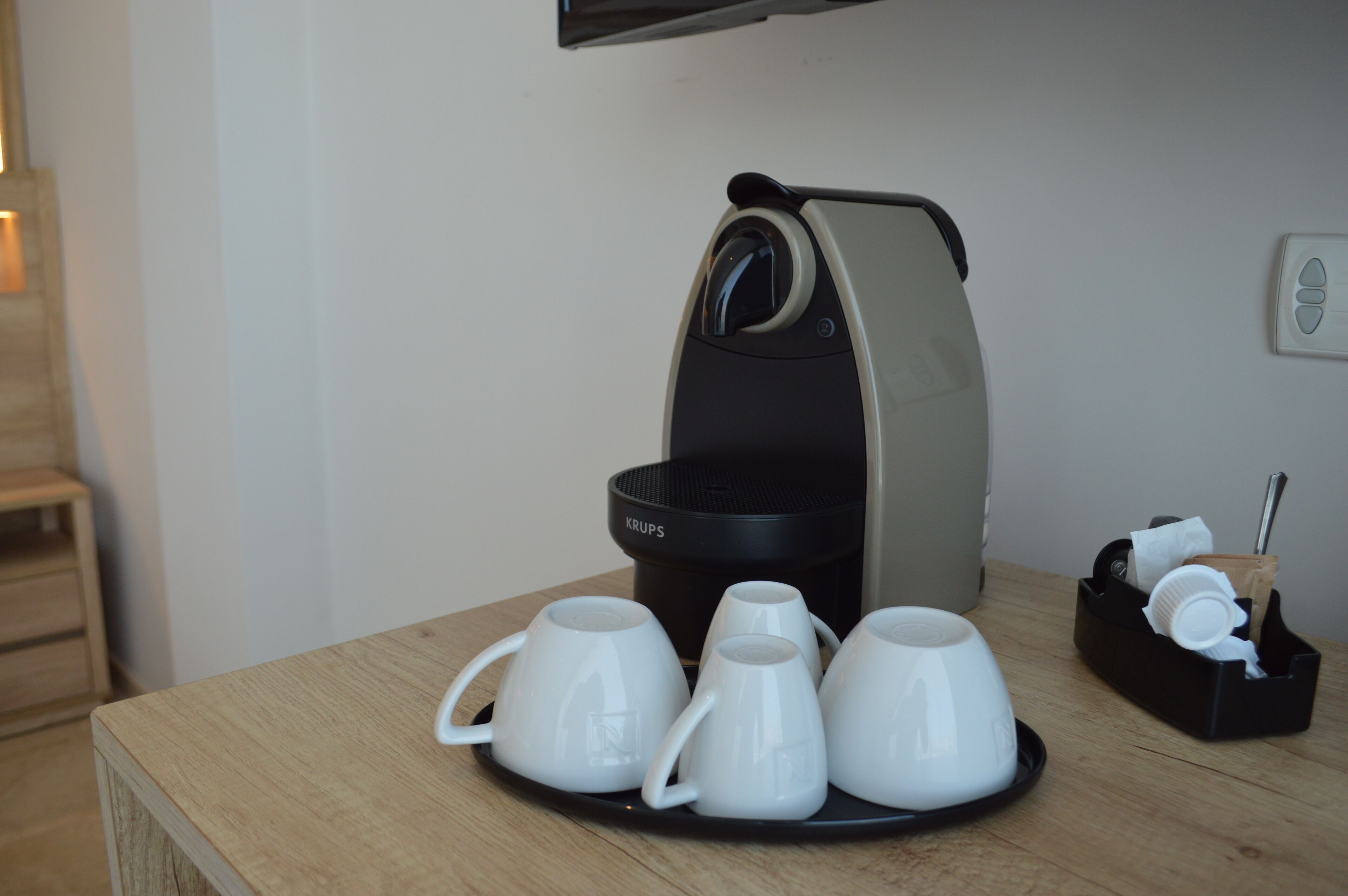 Una cafetera con cuatro tazas blancas sobre una mesa de madera, un recipiente negro con utensilios y un enchufe de pared.