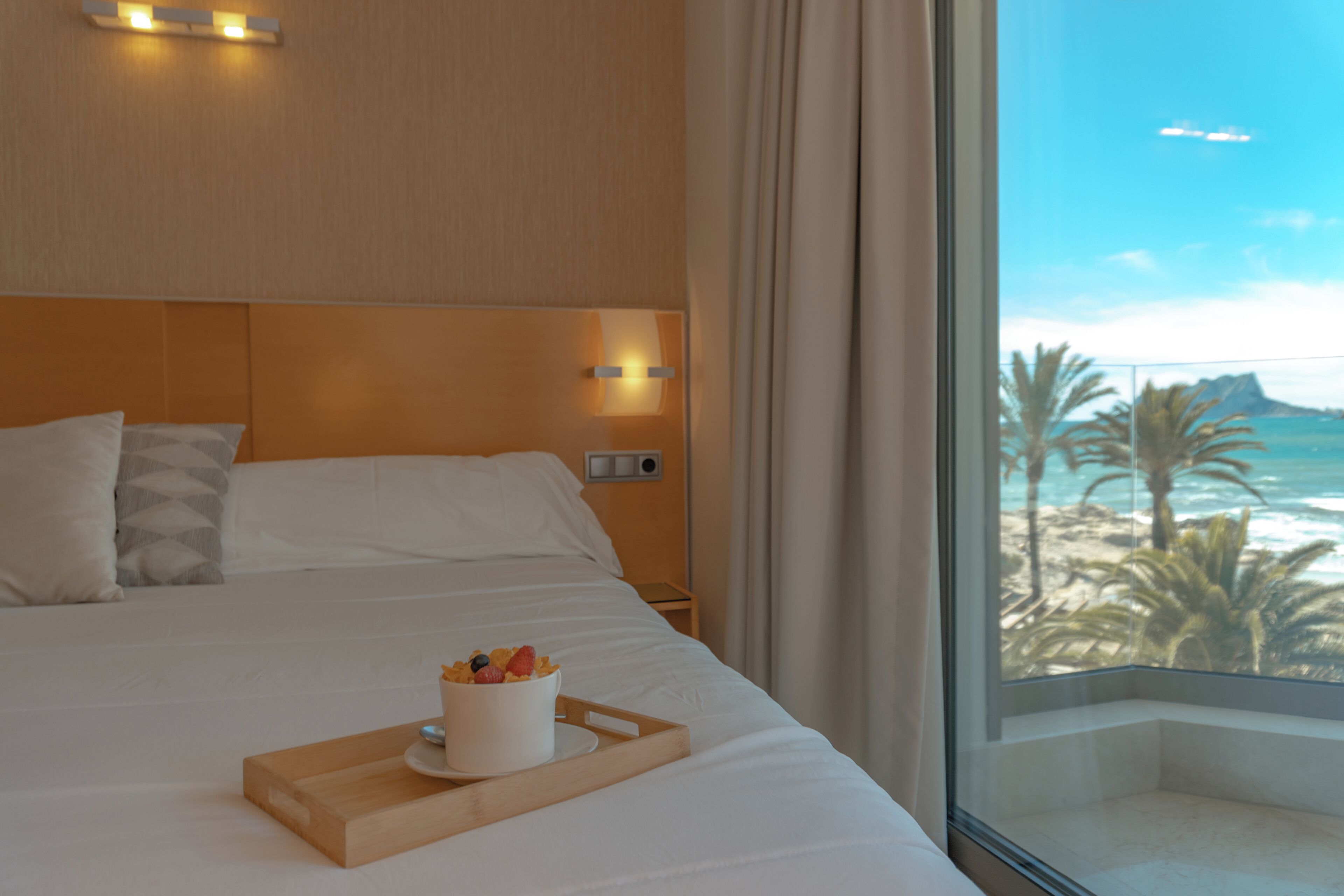 Habitación de hotel con cama, bandeja de frutas, mesita de noche y ventana con vista a la playa y el océano.