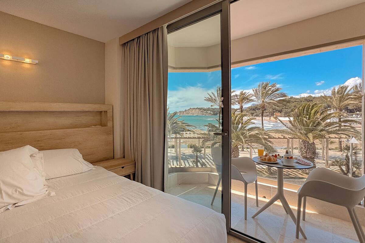 Une chambre d’hôtel dotée d’un grand lit, d’une tête de lit en bois, d’une porte-fenêtre coulissante donnant sur un balcon avec une table et des chaises, et offrant une vue sur la plage et l’océan.