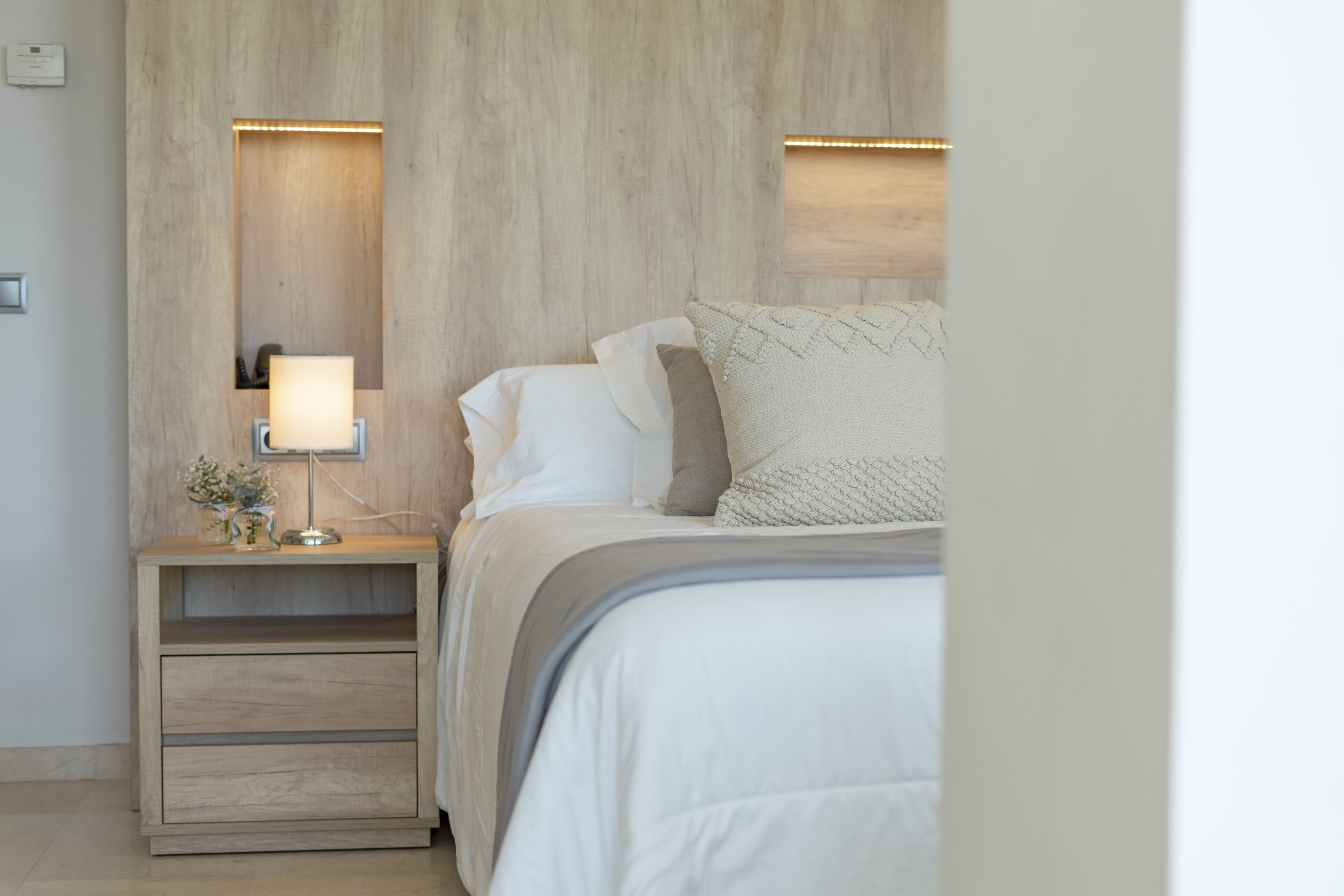 A neatly arranged bedroom with a wooden headboard, a bedside table with a lamp, and a bed with white bedding and decorative pillows.