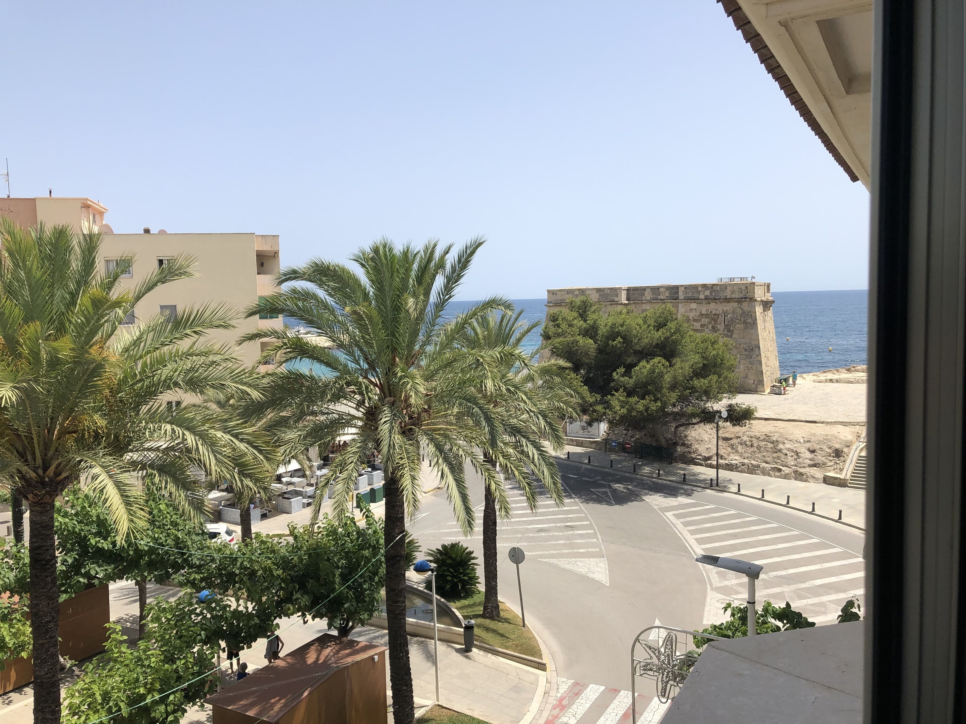 A coastal view with palm trees, a road with pedestrian crossings, and a historic stone building by the sea.