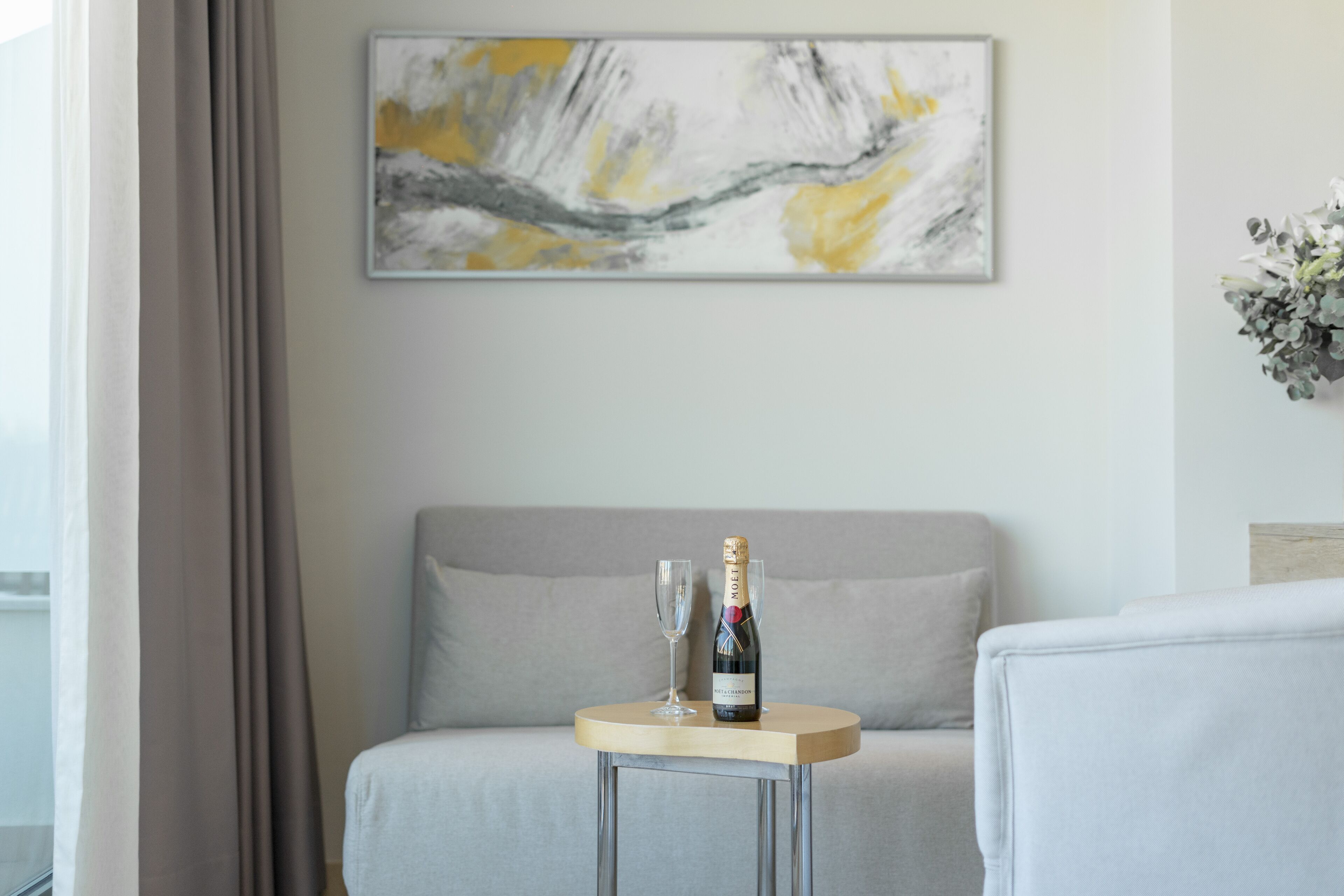 A modern room with a grey sofa, a small round table with a bottle of champagne and a glass, and a large abstract painting on the wall.