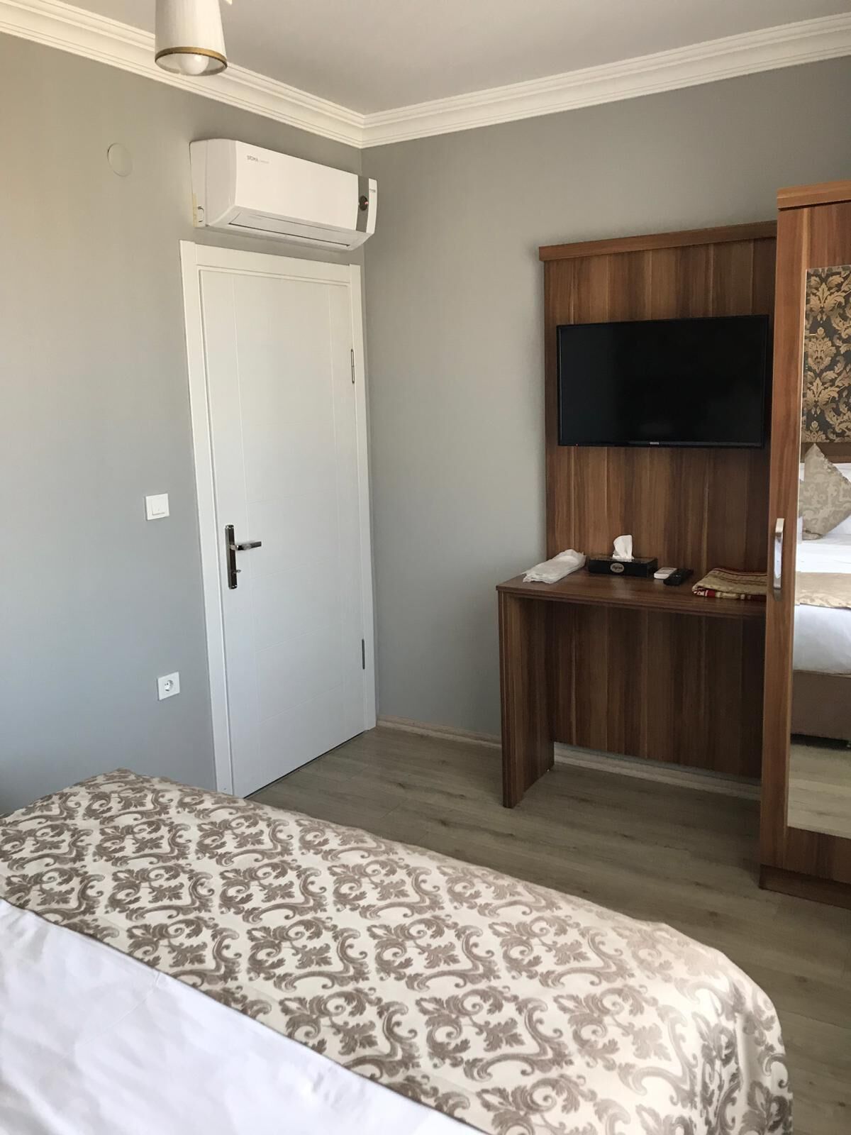 A hotel room with a bed, a desk with a telephone, a flat-screen TV, and an air conditioning unit mounted on the wall.