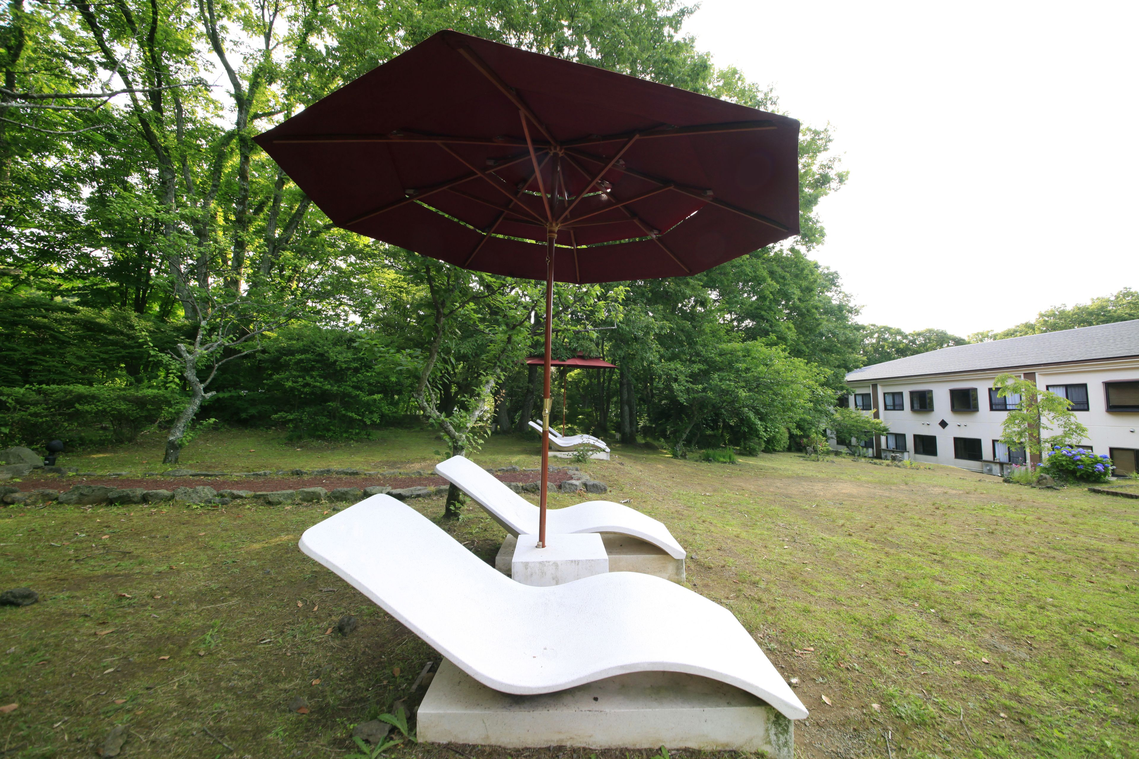 A concrete outdoor seating area with a red umbrella, surrounded by grass and trees.
