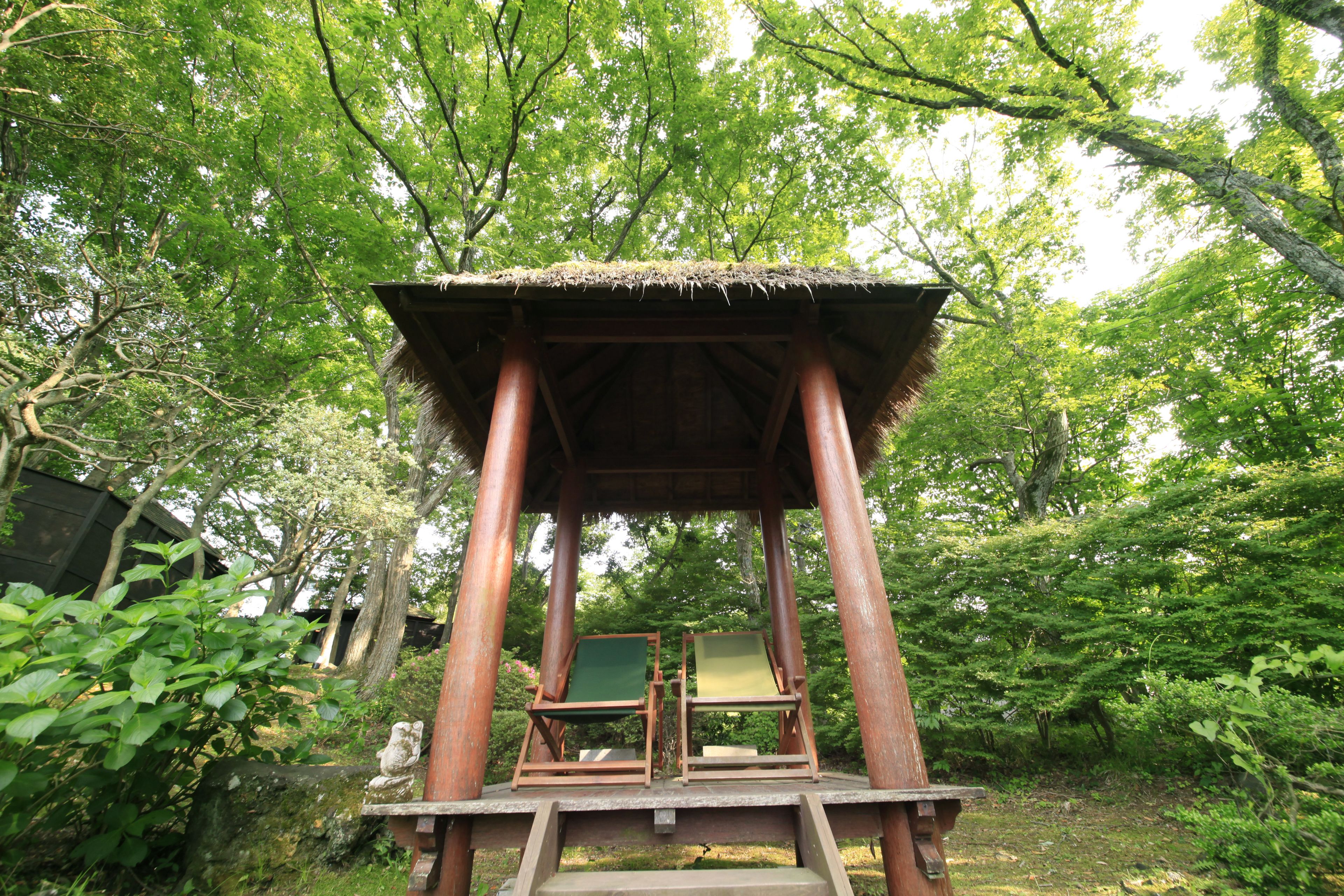 Un kiosque en bois avec un toit de chaume, deux chaises en bois et un banc en pierre.