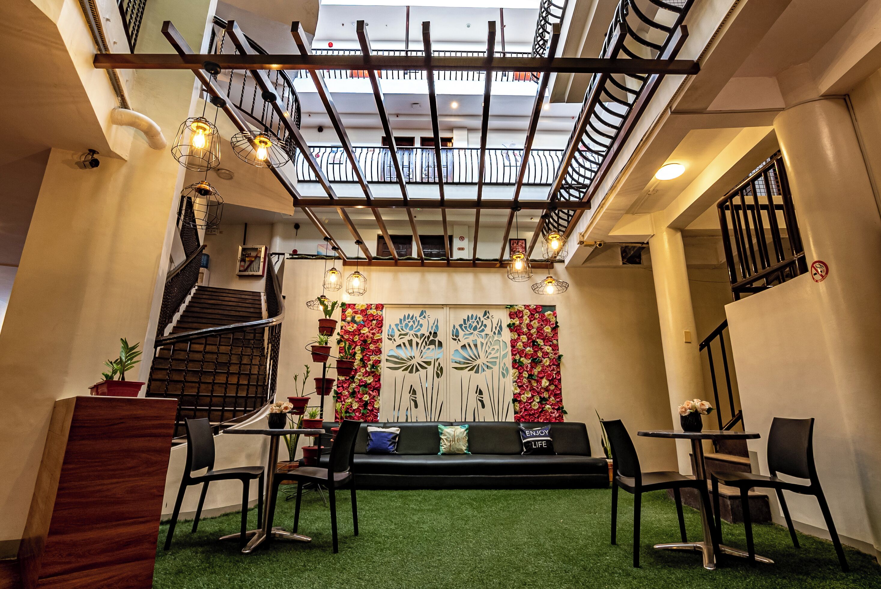 Un espacio interior acogedor con un sofá negro, dos sillas, una mesita con flores, una escalera y una pared decorativa con motivos florales.