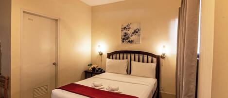 A hotel room with a bed, a ceiling fan, a nightstand with a lamp, and a door.