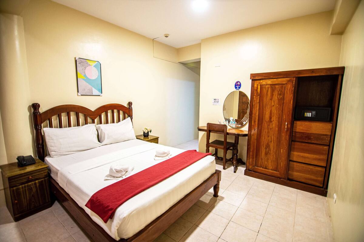A hotel room with a wooden bed, a desk with a chair, a wardrobe, and a small bedside table with a lamp.