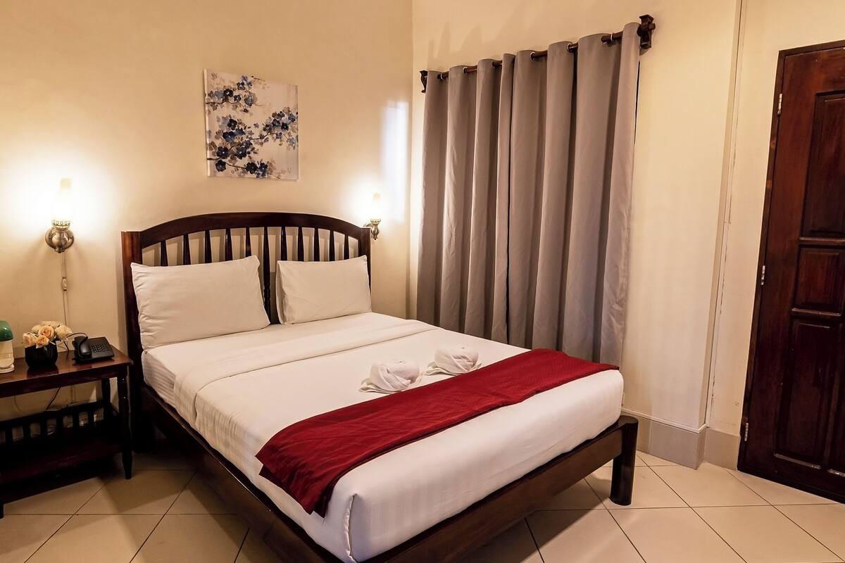 A neatly made bed with white linens and a red throw blanket, a bedside table with a lamp and a phone, and a floral artwork on the wall.