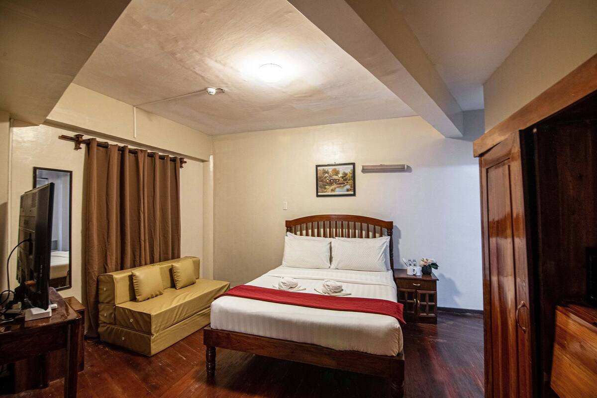 A bedroom with a wooden bed, a beige sofa, a wooden nightstand, and a television on a wooden stand.