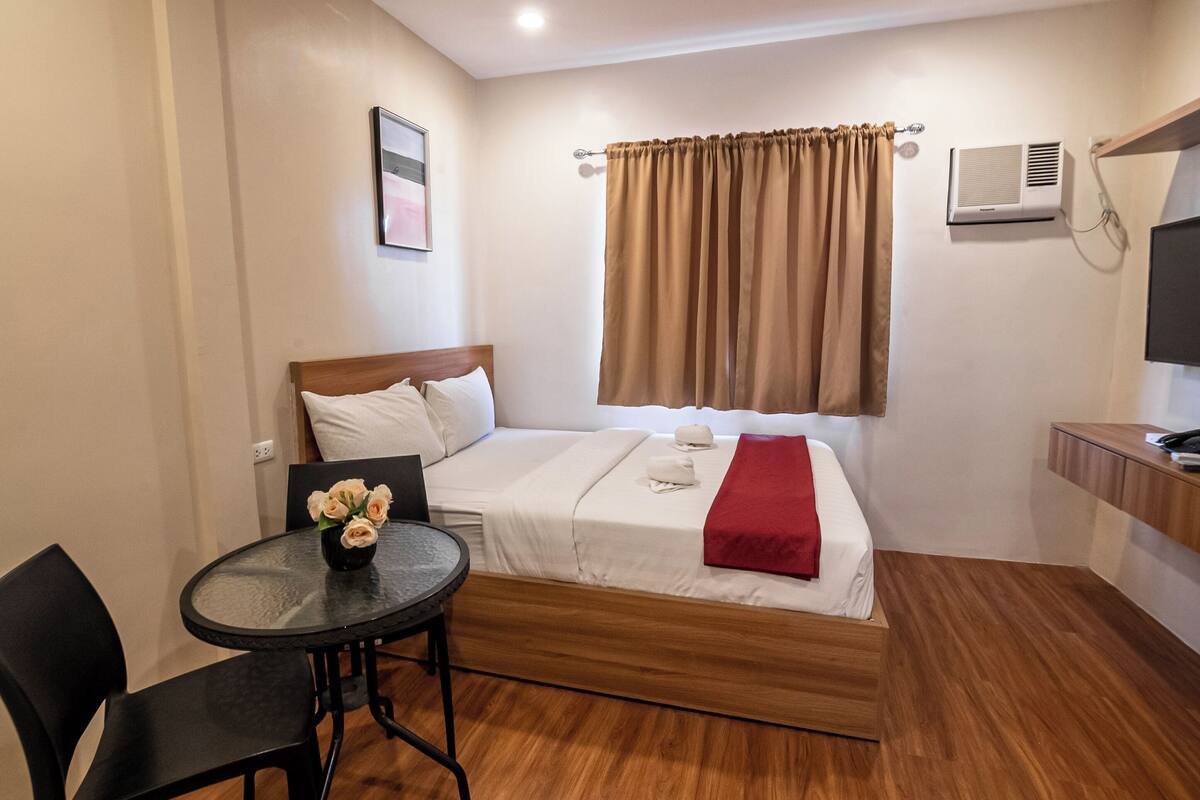 A hotel room with a bed, a small table with chairs, a TV, and an air conditioning unit.