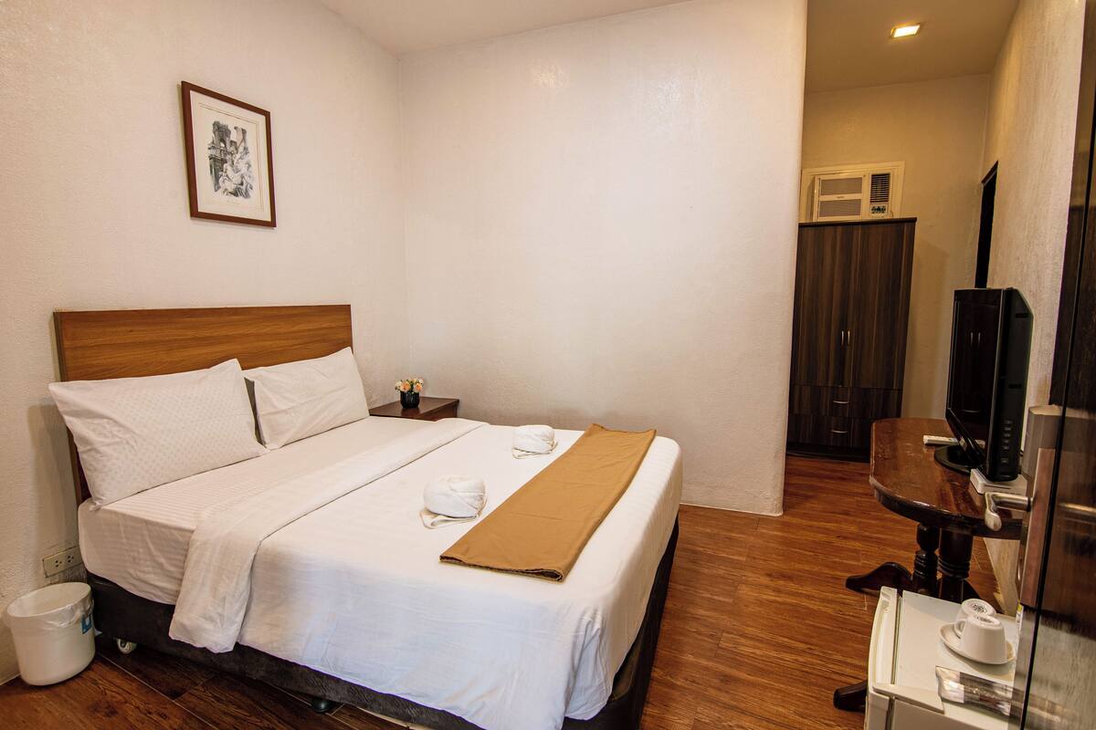 A neatly made bed with white linens and a brown blanket, a small bedside table with a vase of flowers, and a framed picture on the wall.