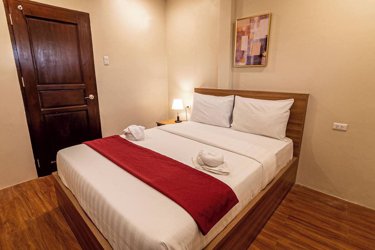 A neatly made bed with white and red bedding, a wooden headboard, and a nightstand with a lamp.