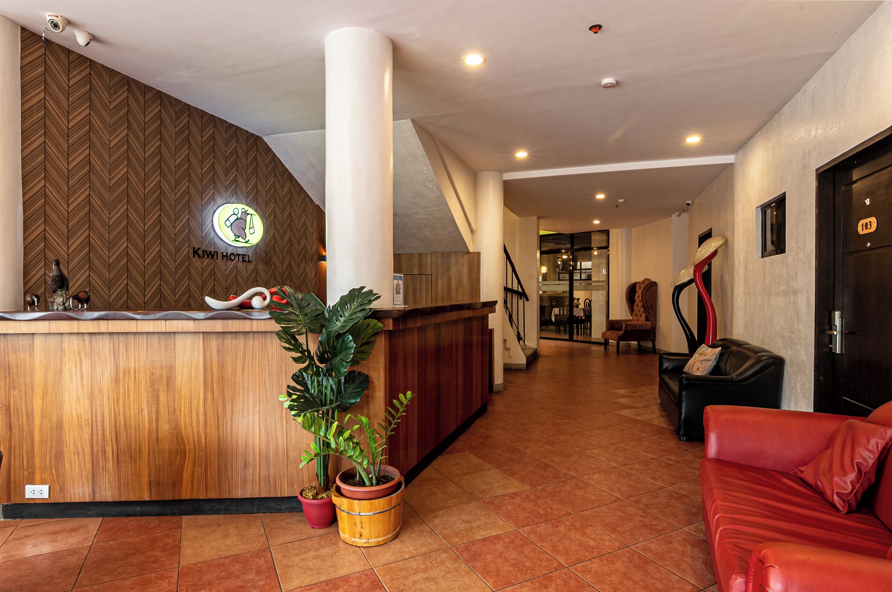 Uma área de recepção de hotel com um balcão de madeira, um vaso de planta, um sofá de couro vermelho e uma escada que leva a outro andar.