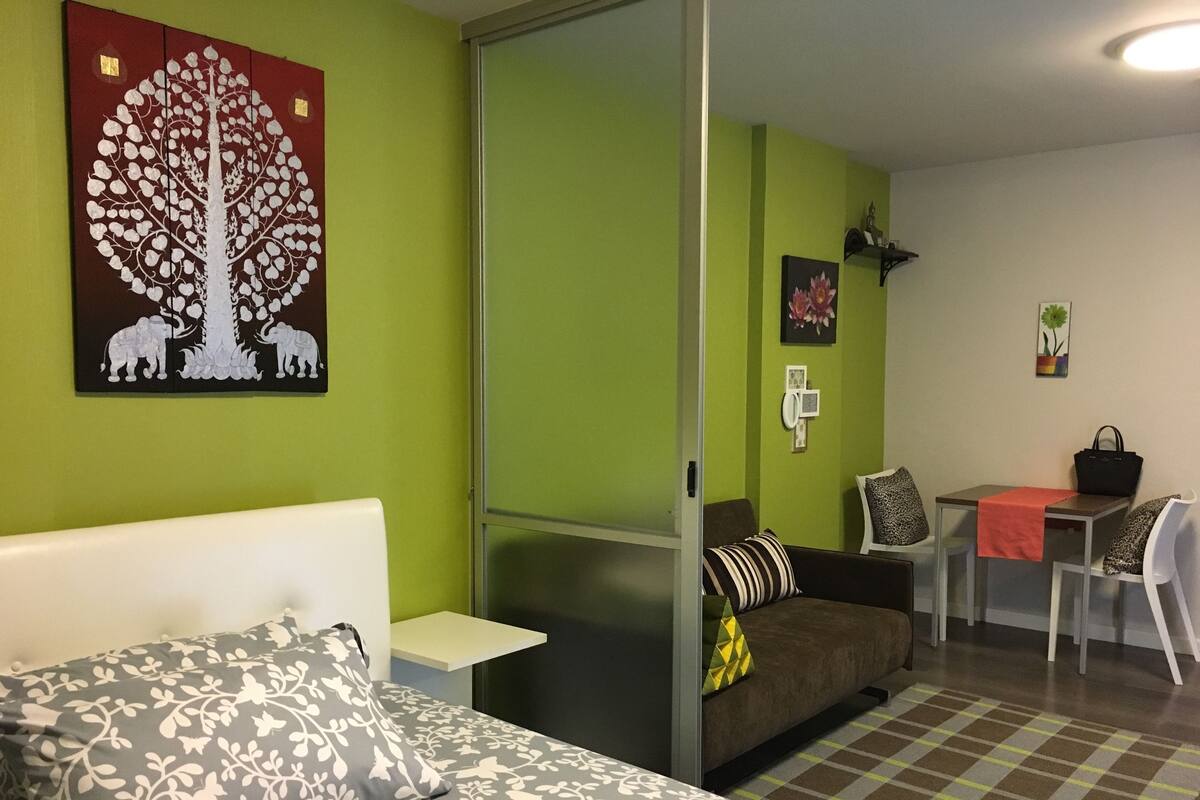 A room with a bed, a sofa, a dining table, and a chair. There is a painting on the wall and a handbag on the table.
