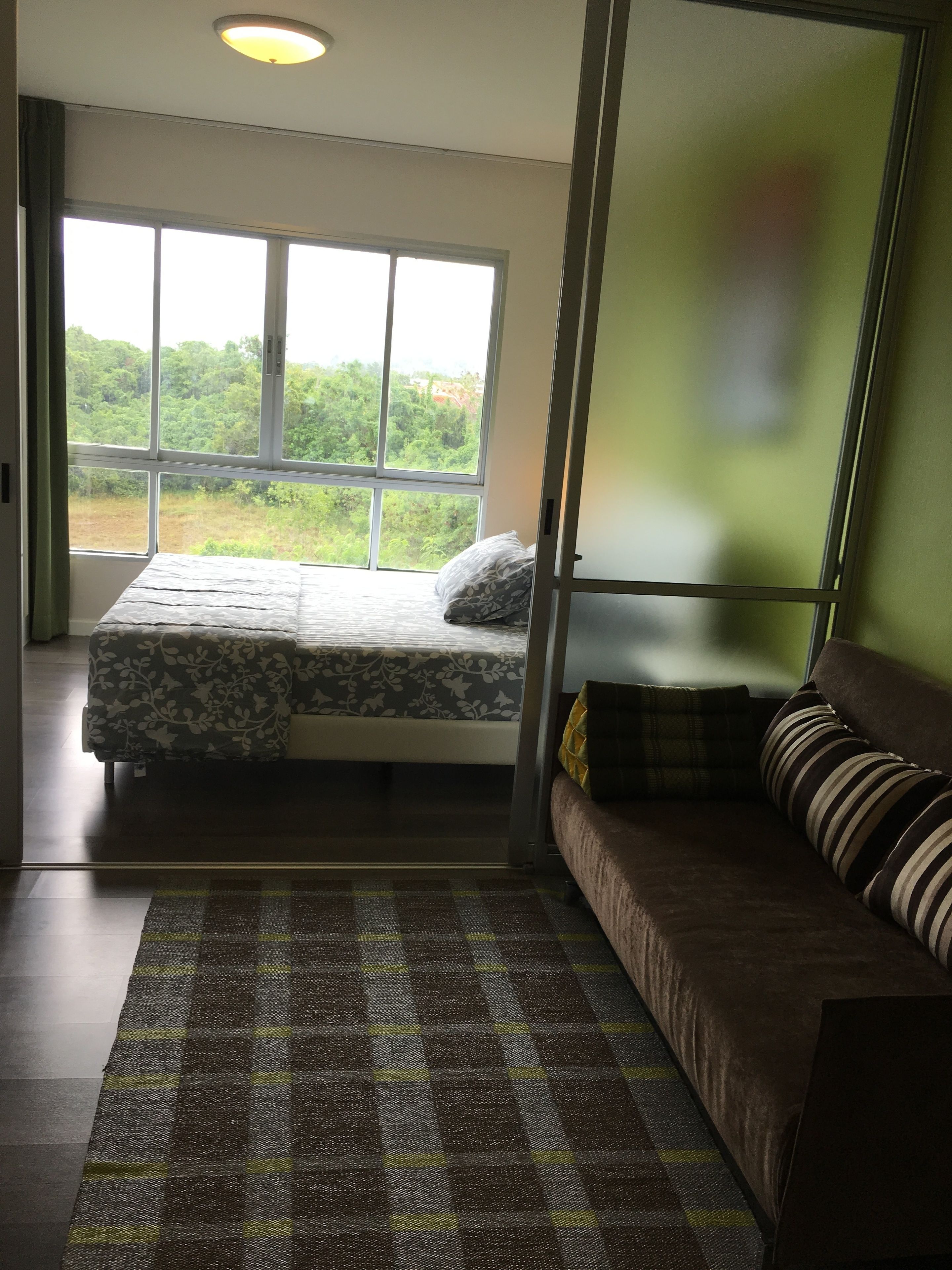 A bedroom with a bed, a sofa, a large window, and a view of greenery.