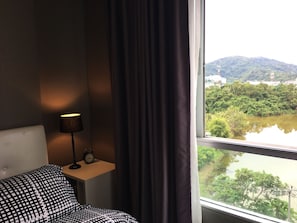A hotel room with a bed, a bedside table with a lamp and an alarm clock, and a window offering a view of a river and greenery. - dcondo mine by BP (Kathu)