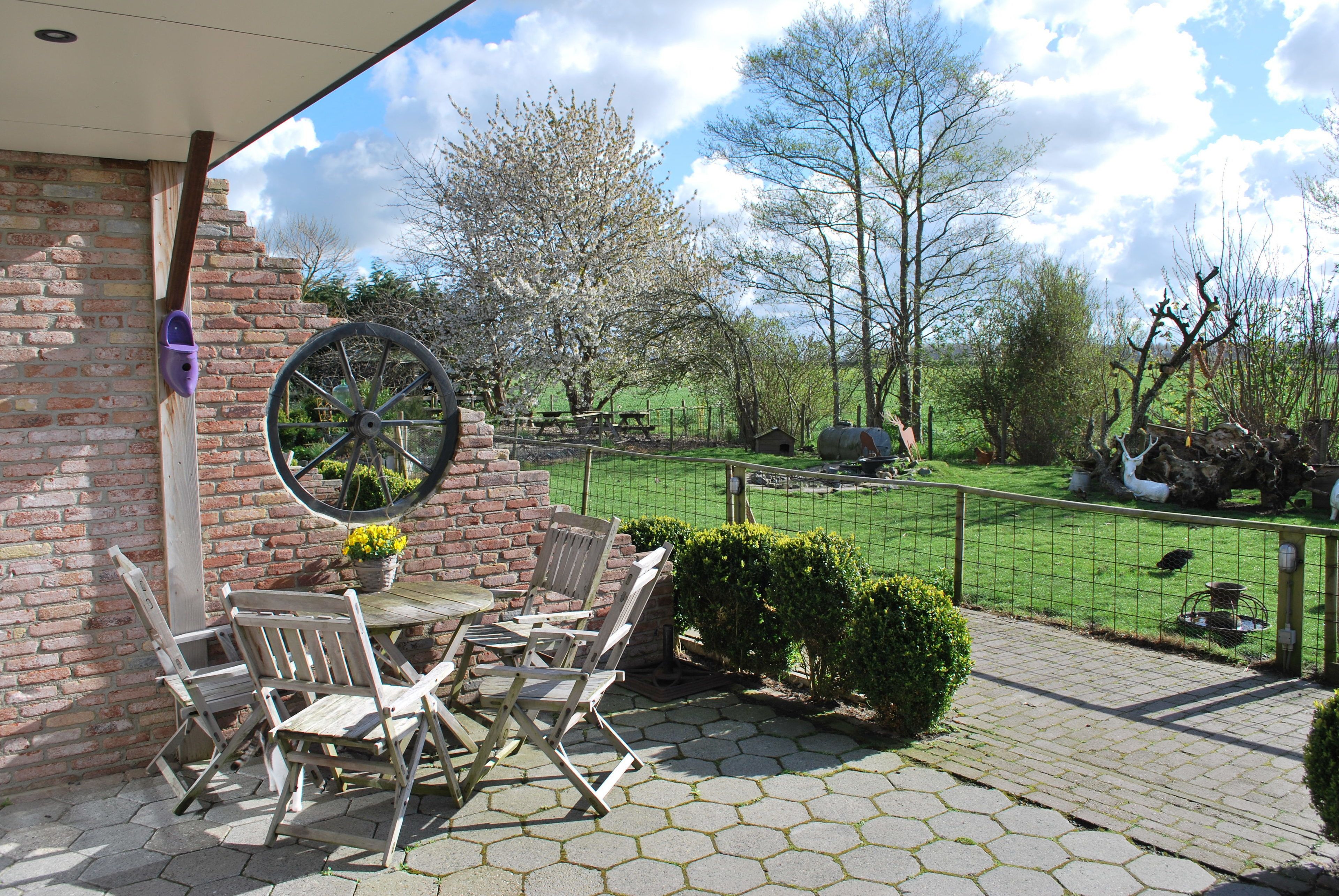 A patio with a brick wall, a wooden table and chairs, a wheelbarrow, and a view of a fenced garden with trees and a picnic table.