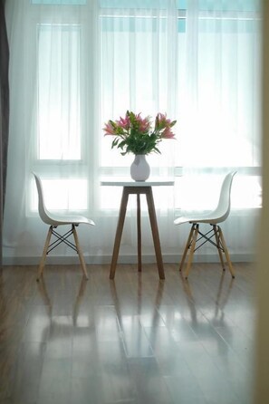 A room with a white table and two white chairs, a vase with flowers, and large windows with sheer curtains. - Rich Hotel (Da Nang)