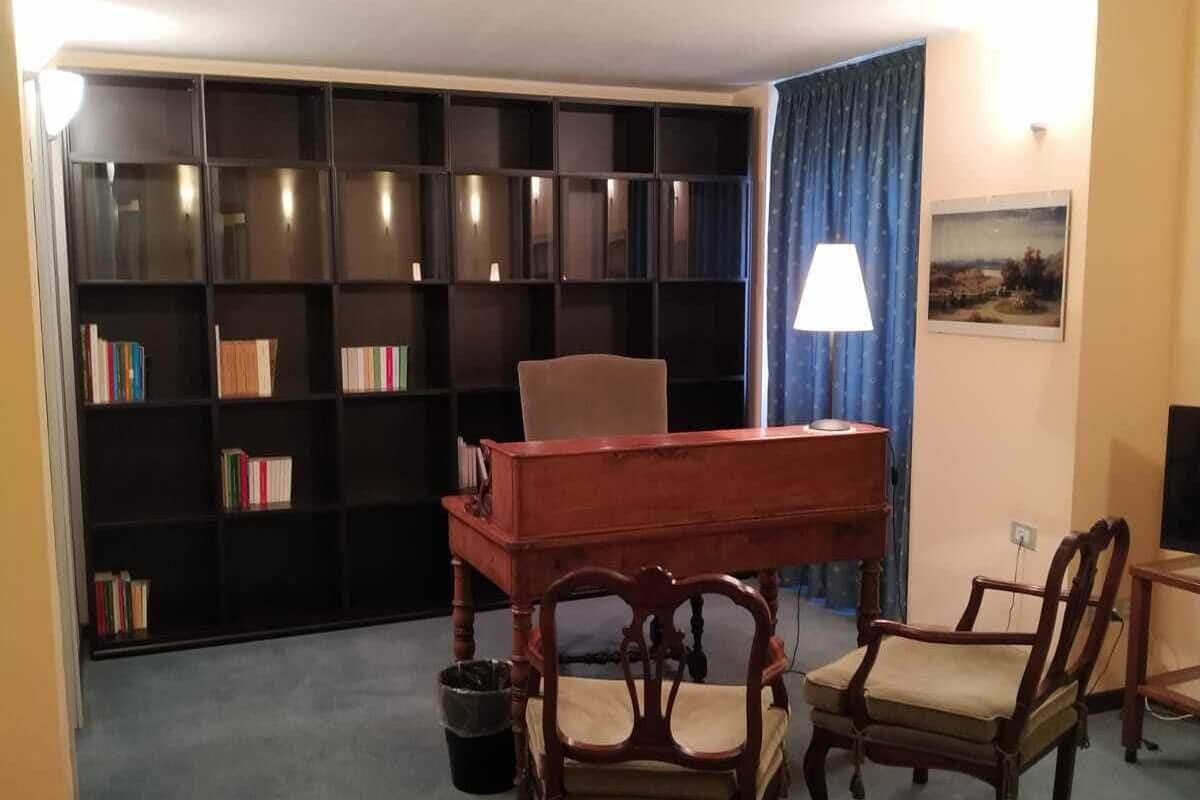 Una habitación con un escritorio de madera, dos sillas, una estantería, una lámpara y un cuadro en la pared.