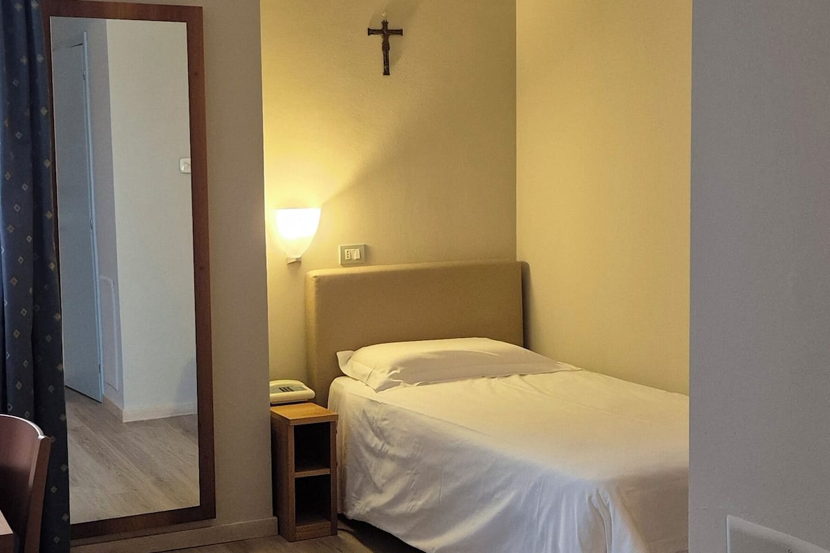 Una habitación de hotel con cama, mesita de noche, lámpara y una cruz en la pared.