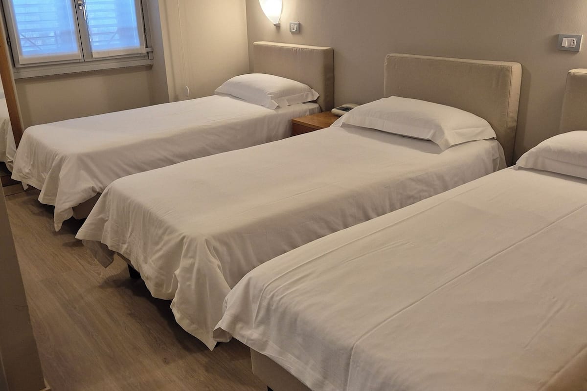Habitación de hotel con tres camas individuales, piso de madera, ventana con persianas y una lámpara fijada a la pared.