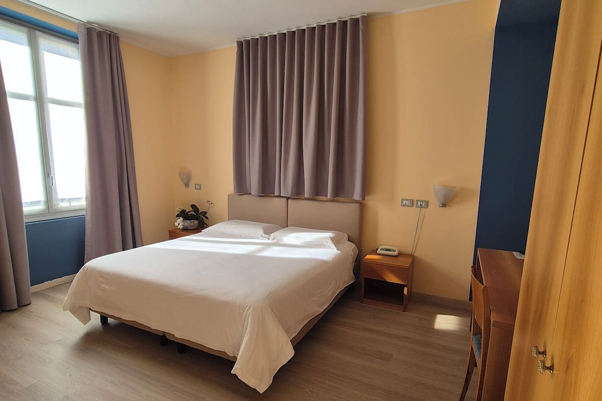 Un dormitorio con una cama grande, una mesita de noche, una lámpara y un ventanal con cortinas.