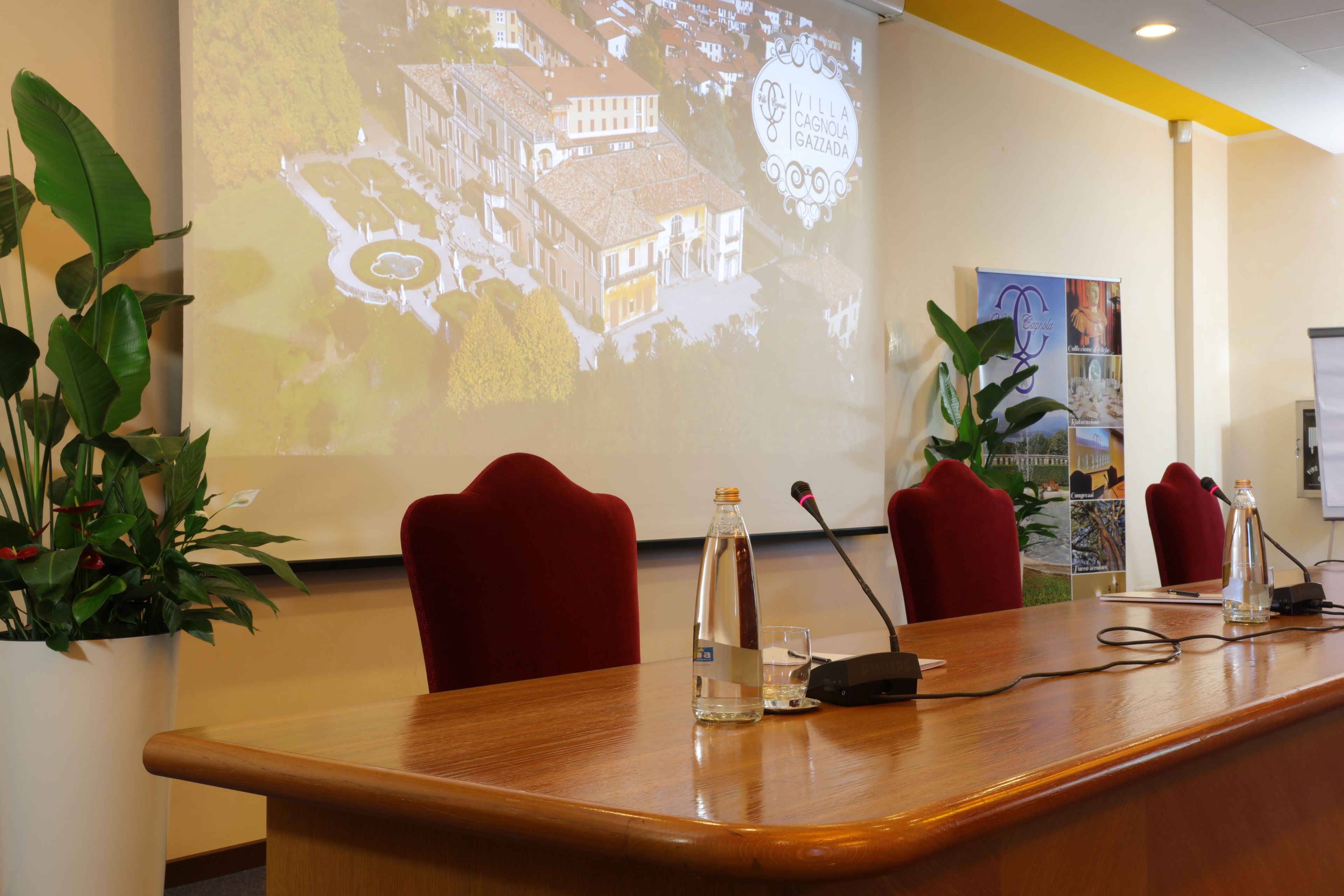 A conference room with a wooden table, red chairs, a projector screen displaying a building image, and a potted plant.