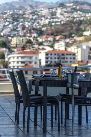 Une terrasse dotée de chaises en osier et d’une table dressée pour le dîner, offrant une vue sur un paysage urbain avec des bâtiments et une montagne en arrière-plan.
