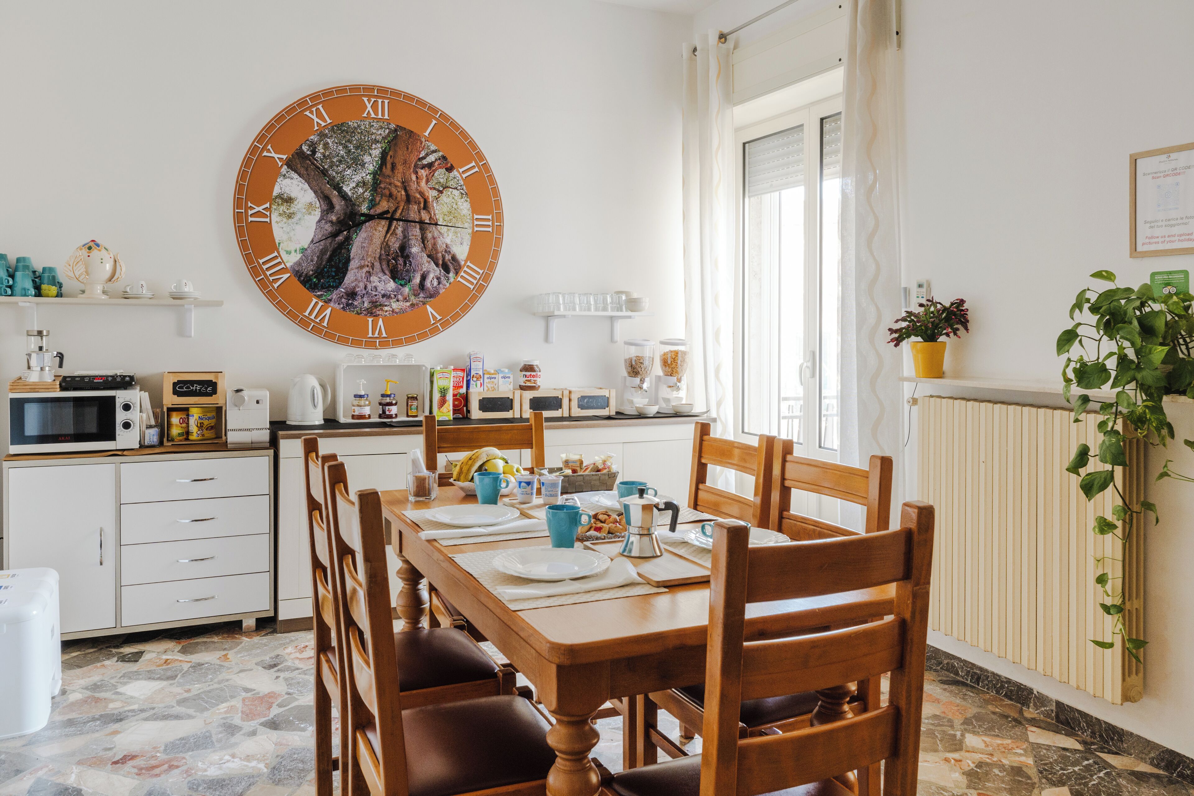 Un coin repas avec une table en bois dressée pour quatre personnes, entourée de chaises. Une grande horloge murale avec des chiffres romains est visible. La pièce a des murs blancs, un sol carrelé et une fenêtre avec des rideaux.
