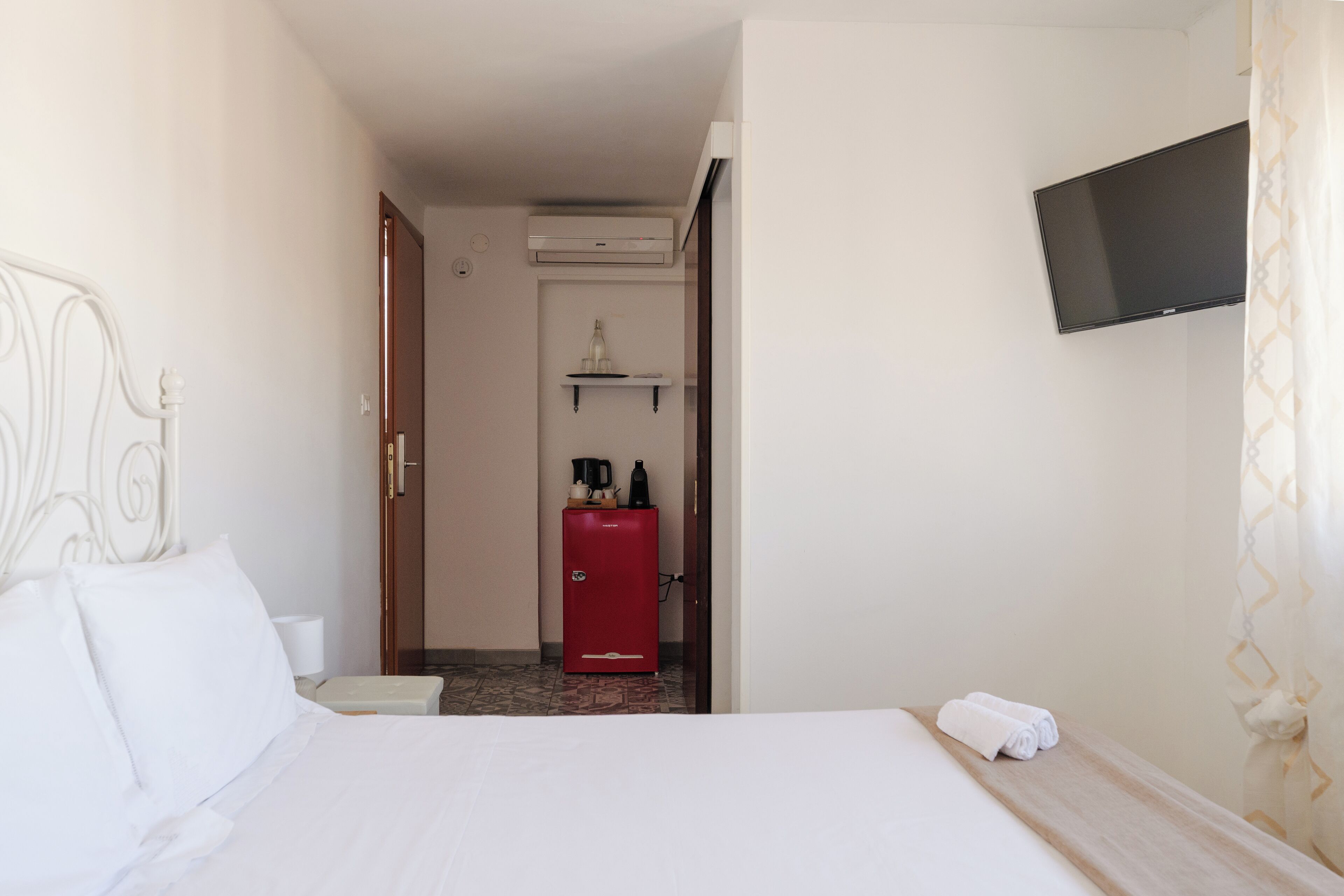 Habitación de hotel con cama, televisor montado en la pared, minibar rojo y una pequeña estantería con hervidor y cafetera.