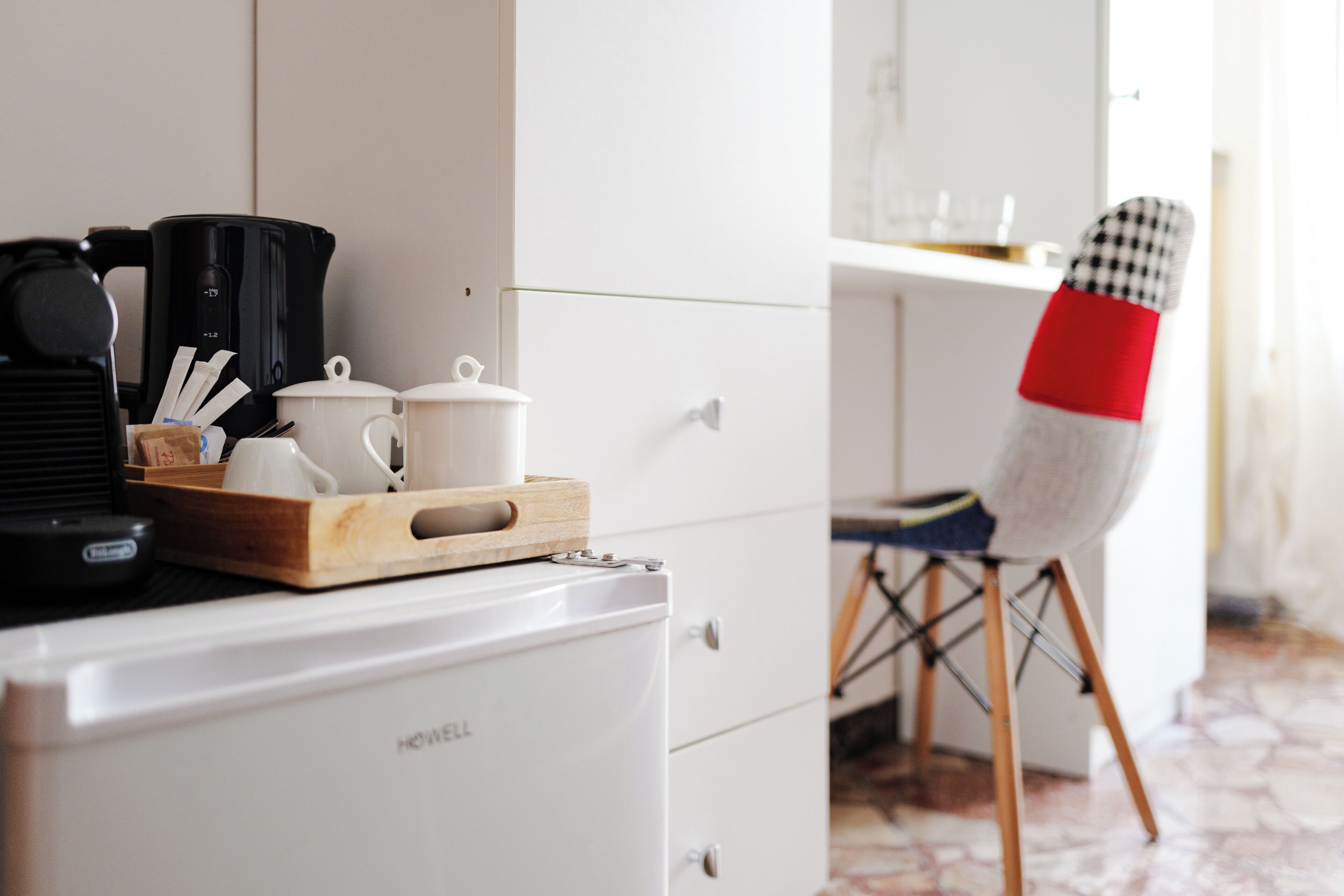 Eine Küchenzeile mit Kaffeemaschine, Mikrowelle und kleinem Kühlschrank. Auf der Arbeitsplatte steht ein Holztablett mit Tassen und einem Teekessel. Ein Stuhl mit rot-weiß kariertem Sitzkissen ist sichtbar.