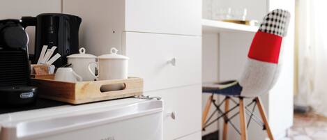 A kitchenette with a coffee maker, a microwave, and a small refrigerator. There is a wooden tray with cups and a teapot on the counter. A chair with a red and white checkered cushion is visible.