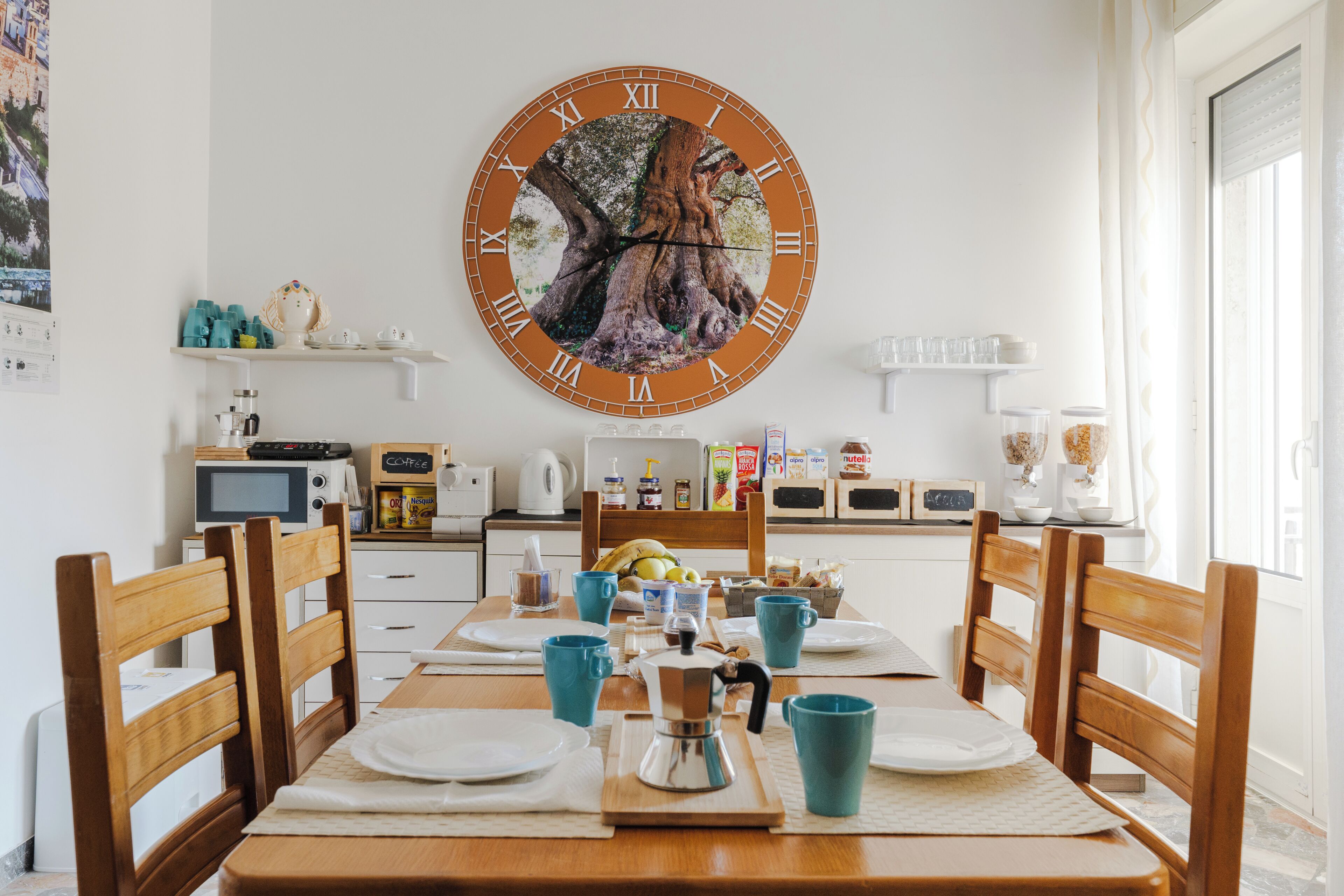 Een eetruimte met een houten tafel gedekt voor vier personen, een grote wandklok en een keuken op de achtergrond.