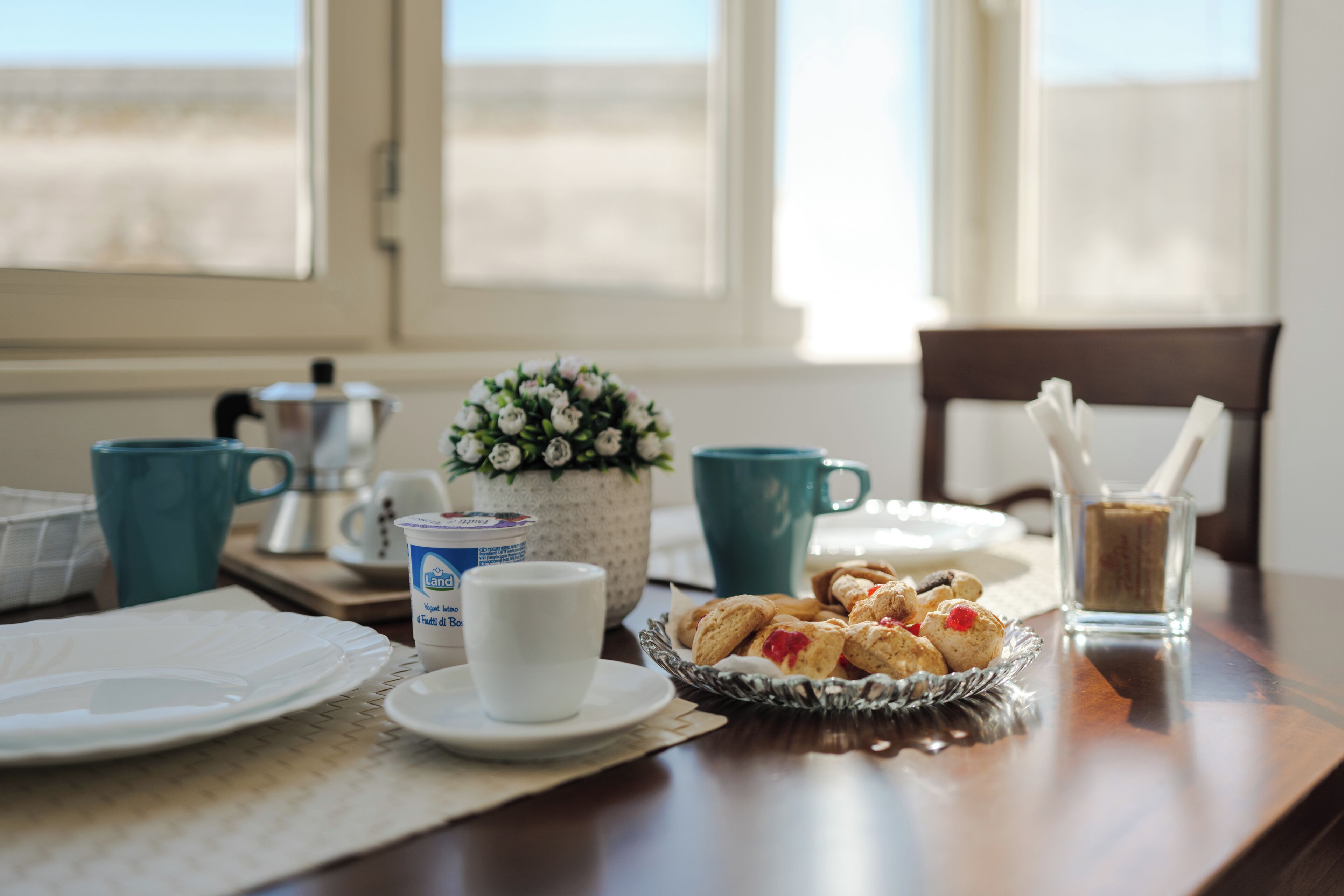 Une table de petit-déjeuner avec une cafetière, des tasses, un vase de fleurs et des viennoiseries.