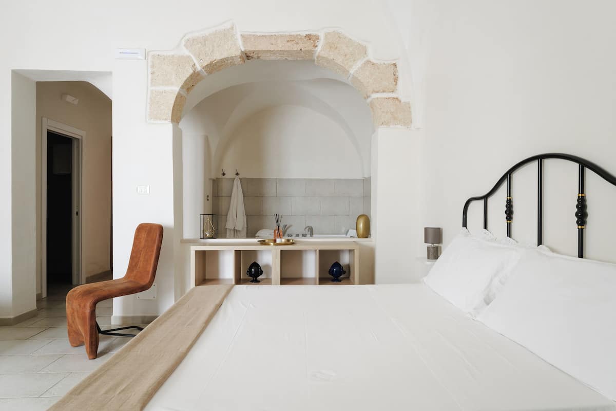 Un dormitorio con cama, una silla de madera, una tocador con espejo y un arco de piedra.