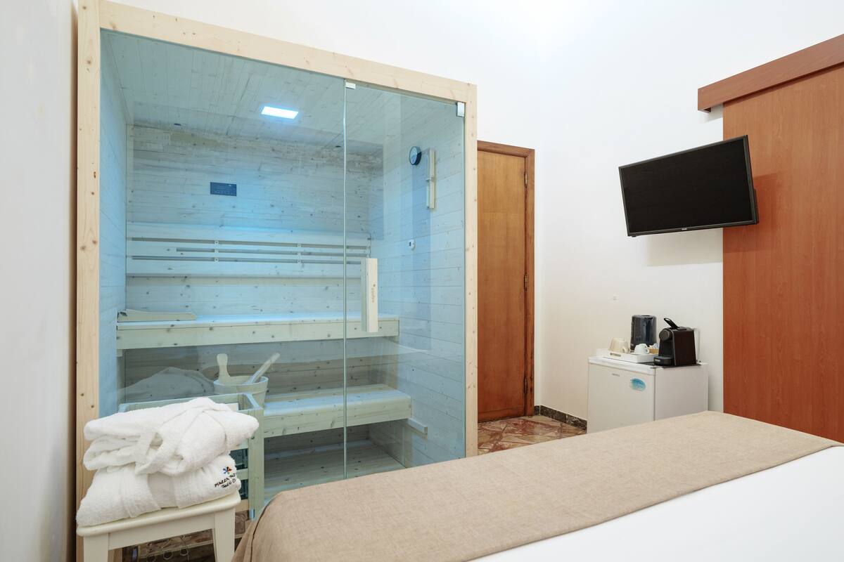 Habitación de hotel con sauna acristalada, cama, escritorio pequeño con cafetera y televisor de pantalla plana montado en la pared.
