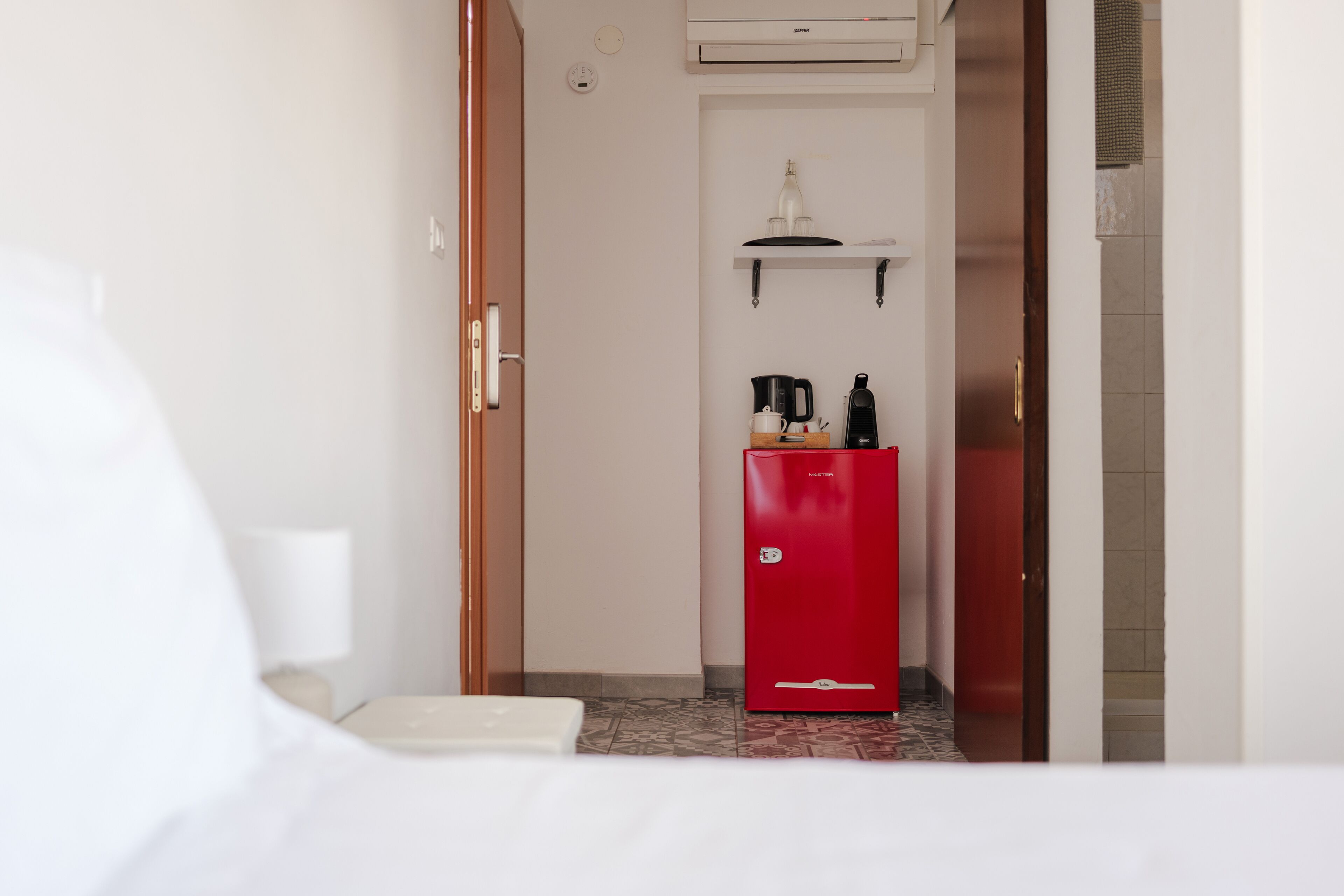 A hotel room with a bed, a small kitchenette with a red refrigerator, a coffee maker, and a shelf with bottles.