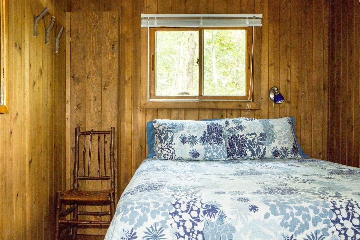A wooden cabin room with a bed, a chair, a window, and a lamp.