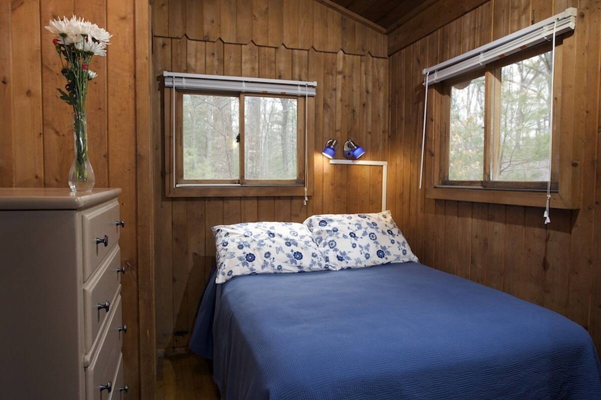 A wooden cabin bedroom with a bed, dresser, and two windows with blinds.