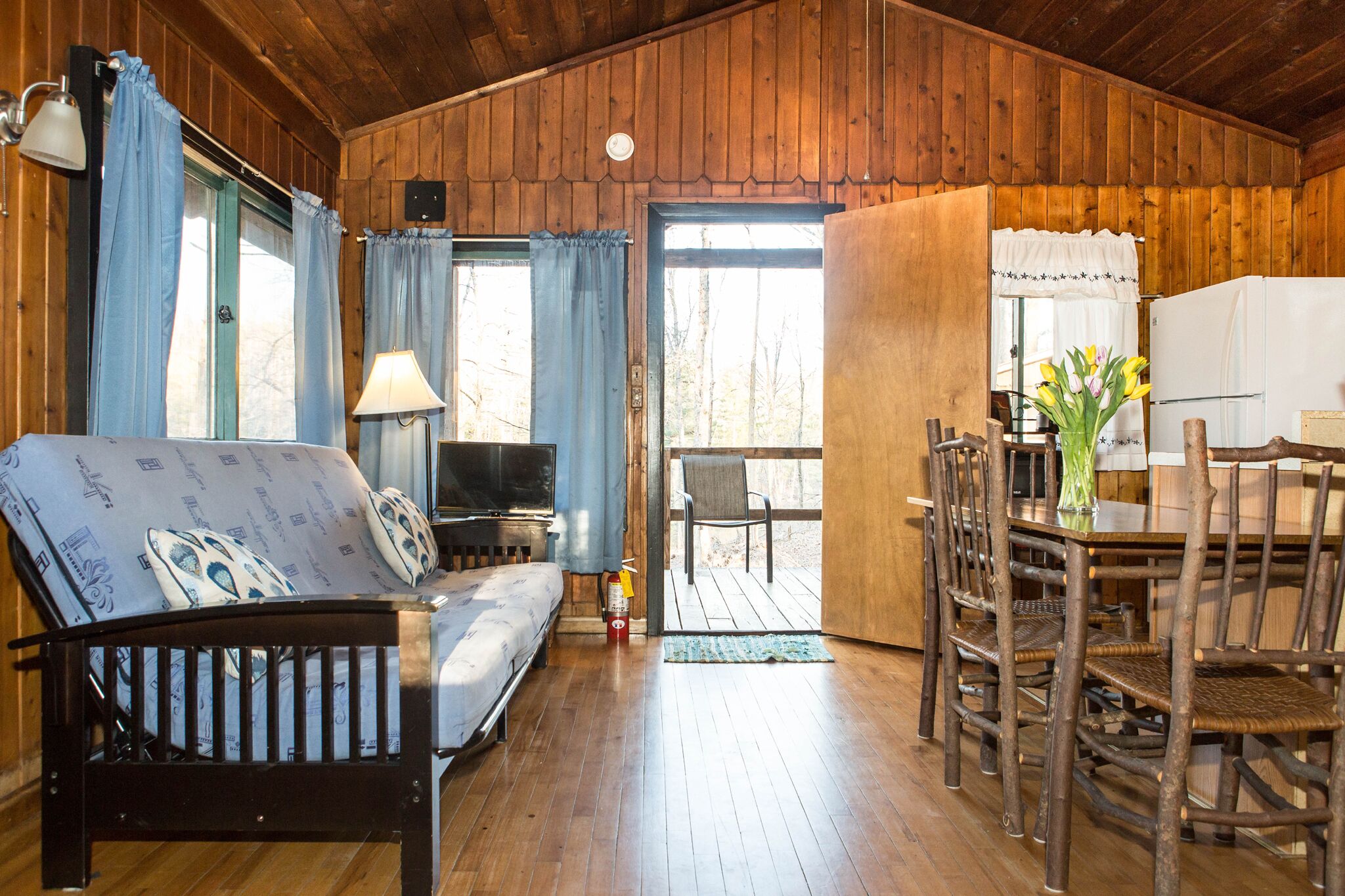 Interior acolhedor de uma cabana com decoração em madeira, um sofá, uma mesa de jantar com cadeiras e uma pequena cozinha.