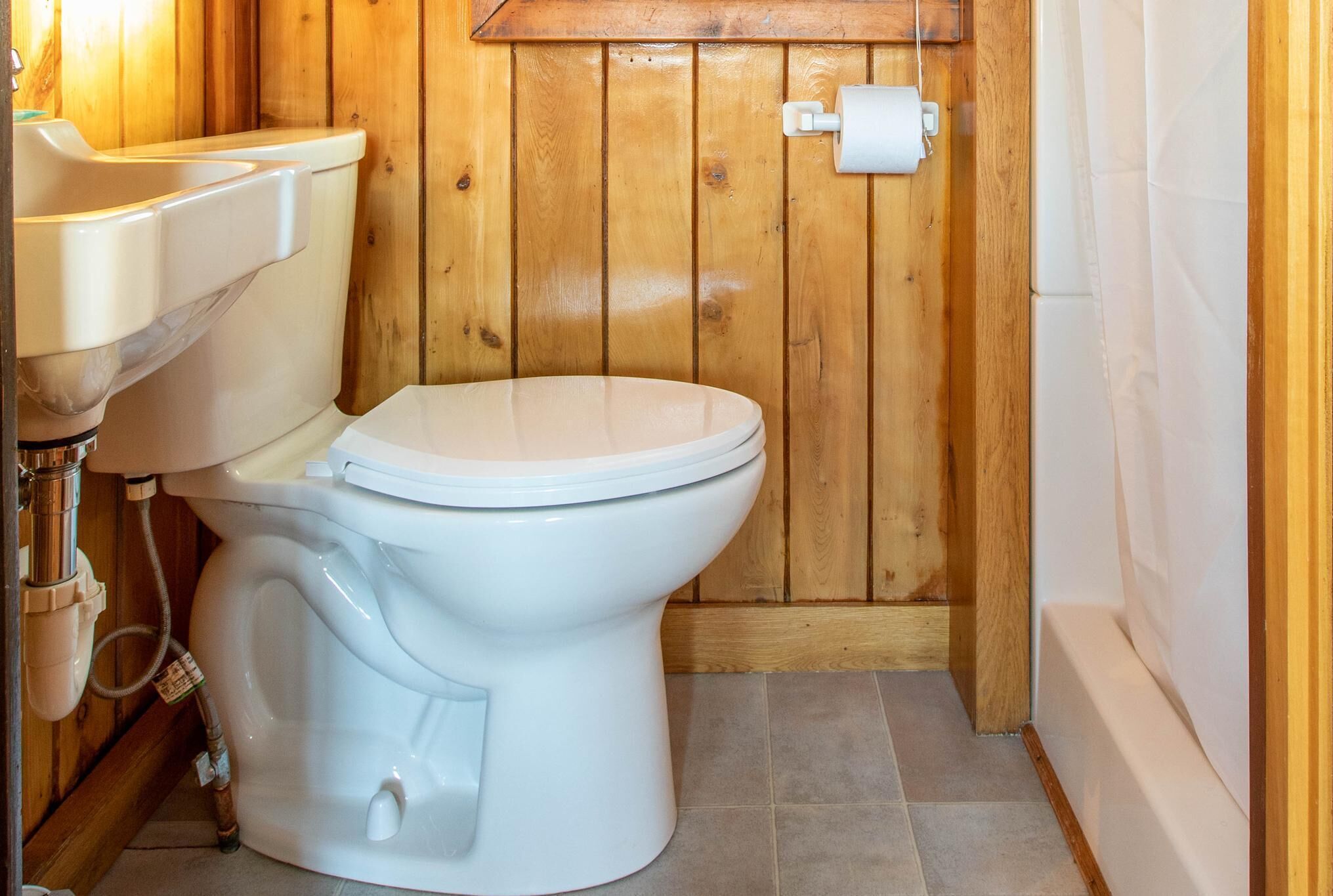 A small, wooden-paneled bathroom with a toilet, sink, and shower.