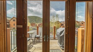 Eine Holzterrasse mit Tisch und Stühlen, ein überdachter Grill und der Blick auf Häuser und Berge durch eine Glastür.
